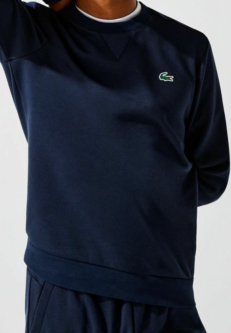 Personne portant un sweat-shirt à manches longues bleu foncé avec un petit logo crocodile vert sur la poitrine, debout devant un fond uni.