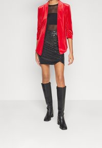 Femme portant des bottes hautes noires, une mini-jupe noire à sequins, un haut noir transparent et un blazer en velours rouge, debout sur un sol blanc uni.