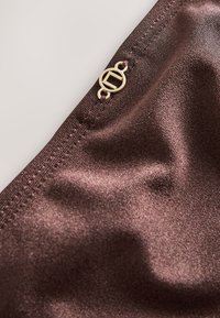 Gros plan sur un tissu marron lisse avec une couture soignée et un petit logo métallique rond doré attaché près de la couture.