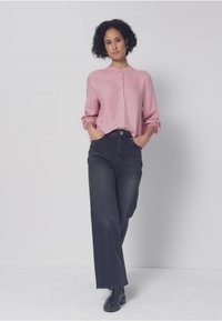 Koszula na guziki z długim rękawem w miękkim różowym materiale, zestawiona z ciemnymi dżinsami o wysokim stanie. Modelka ma na sobie czarne botki za kostkę.