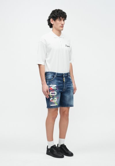 Short en jean avec éclaboussures de peinture multicolores et texte "NO" d'un côté, associé à un polo blanc et des baskets noires.