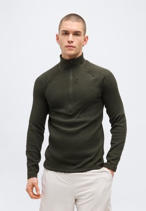 Άνδρας με κοντά μαλλιά που φοράει σκούρο πράσινο πλεκτό πουλόβερ με φερμουάρ στο λαιμό και ανοιχτόχρωμο μπεζ παντελόνι, στέκεται με το ένα χέρι στην τσέπη.