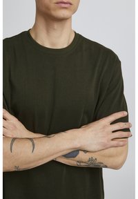 Mørkegrøn bomulds t-shirt med rund hals, korte ærmer og en glat tekstur. Armene er korslagte, hvilket viser forskellige tatoveringer på underarmene.