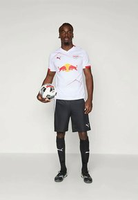 Biely športový dres s červenými akcentmi a logom červeného býka, skombinovaný s čiernymi šortkami a ponožkami, drží vzorovanú futbalovú loptu.