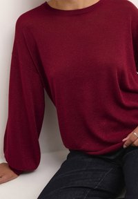 Pull en maille bordeaux avec manches longues légèrement bouffantes et col rond. Texture fine et scintillante ajoutant du détail. Associé à un jean foncé.