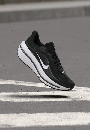 Zapatilla de correr Nike en blanco y negro en el aire sobre asfalto con marcas blancas de calle y pequeños escombros dispersos.