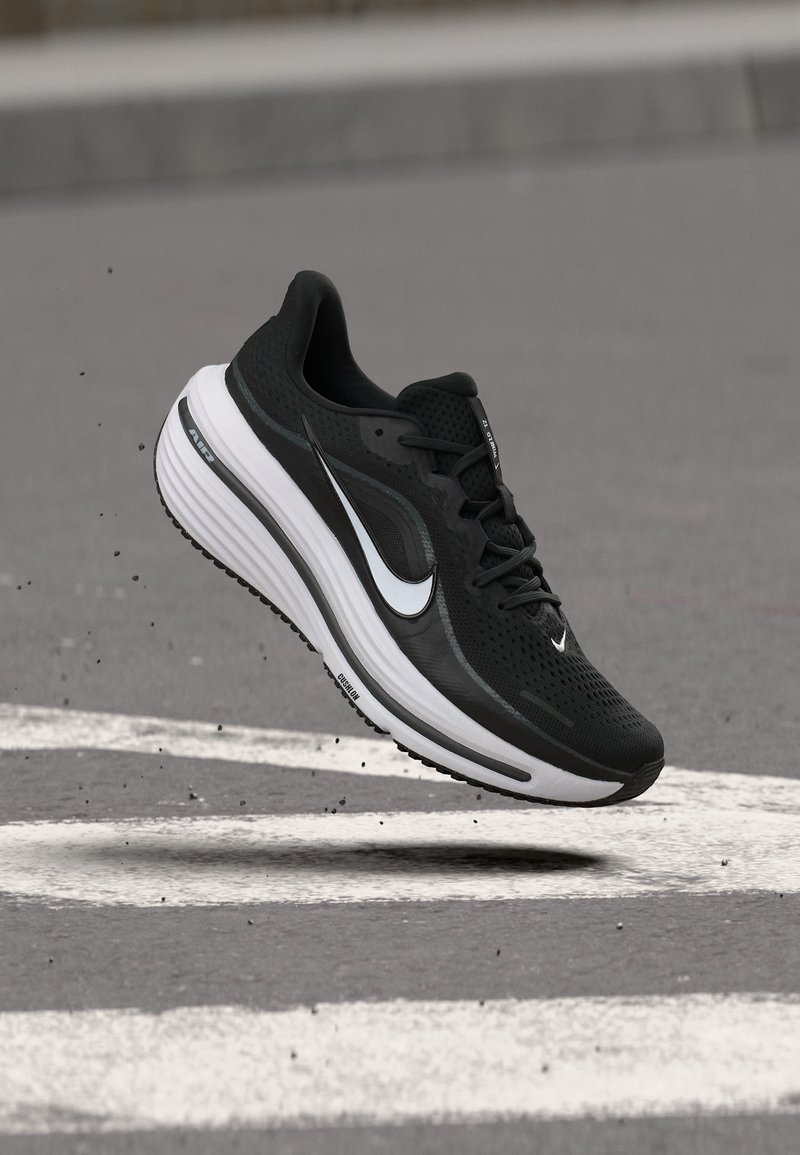Chaussure de course Nike noir et blanc en l'air au-dessus de l'asphalte avec des marquages blancs et de petits débris éparpillés.