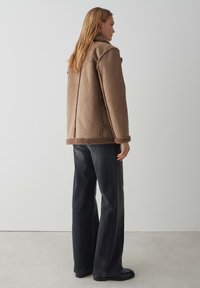 Bruine schapenvachtjas met een fluffy kraag en zoom, gecombineerd met donkere wijde jeans. De jas heeft zichtbare stikdetails.