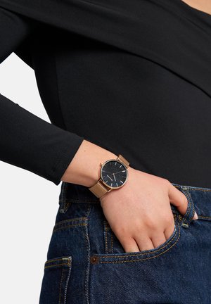 Rund ur med sort urskive, guld mesh-rem og minimalistisk design. Båret på et håndled med denimjeans synlige i baggrunden.