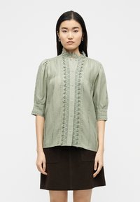 Γυναίκα που φοράει μια ανοιχτό πράσινη μπλούζα με δαντέλα στα κουμπιά και φουσκωτά μανίκια, καθώς και μια σκοτεινή καφέ φούστα, στέκεται μπροστά σε απλό λευκό φόντο.