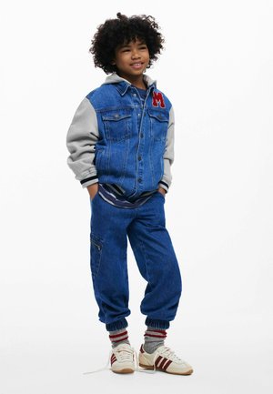 Denim jacket with gray sleeves, embroidered "M" on chest. Blue jeans with elastic cuffs, paired with white sneakers and striped socks.