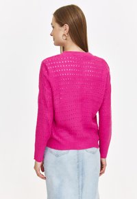 Różowy sweter z dzianiny o ażurowym wzorze, z długimi rękawami i ściągaczem u dołu. Nosi się go z jasnoniebieską spódnicą dżinsową.