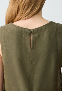 Top in lino verde oliva con scollatura rotonda, caratterizzato da una piccola chiusura a bottoncino e uno spacco sul retro. Tessuto leggero e testurizzato.
