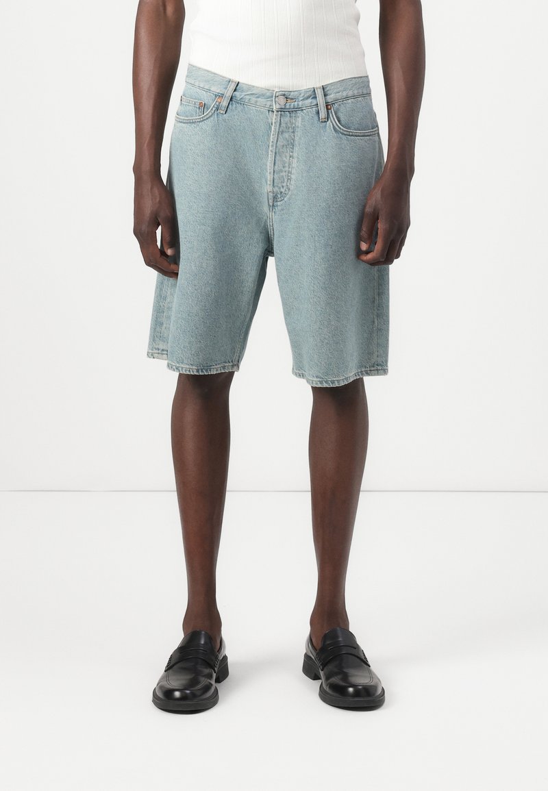 Shorts en denim bleu clair avec une coupe décontractée, dotés d'une taille haute, d'un design à cinq poches et d'un ourlet brut. Accompagnés de chaussures noires à enfiler.