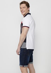 Polo din bumbac cu mânecă scurtă pentru bărbați, având material alb, guler bleumarin, accente roșii și logo. Asoclat cu pantaloni scurți din denim închis la culoare, cu manșete răsucite.