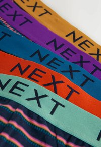 Ceintures élastiques colorées en moutarde, violet, bleu, orange et sarcelle avec le mot « NEXT » imprimé en lettres noires.