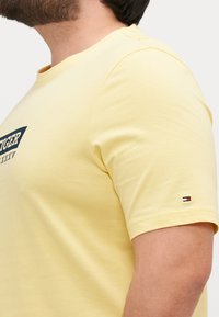 Gul bomulls T-skjorte med rund hals, korte ermer, og et marineblått logo print på fronten. Har en liten tri-farge aksent på ermet.