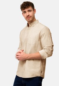 Beige Langarmhemd mit Button-Down-Kragen, einer einzelnen Brusttasche und hochgerollten Ärmeln. Es zeichnet sich durch einen strukturierten Stoff und ein seitliches Etikettendetail aus.
