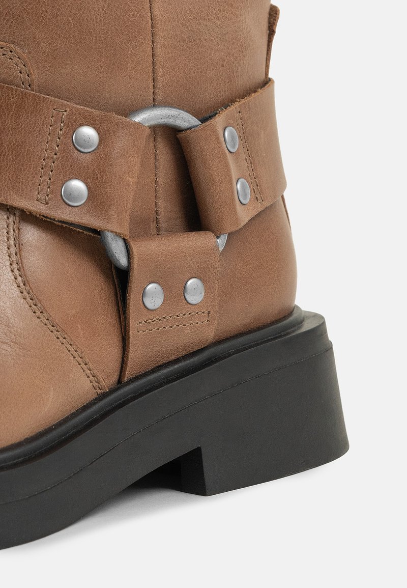 Botín de piel marrón hasta el tobillo con punta redonda, acentos de hardware plateado y un tacón negro grueso con una suela texturizada.