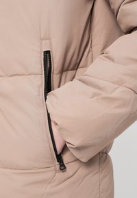 Mano con manguito de chaqueta acolchada beige, con los dedos dentro de un bolsillo con cremallera negro parcialmente abierto.