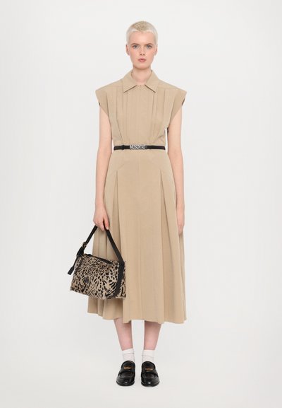 Beige pleated dress with a collar, belted at the waist. The model holds a leopard-print handbag, wears black loafers, and white socks.