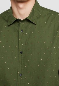 Grüne Hemdbluse mit einem Rautenmuster in Hellbraun, klassischem Kragen und schwarzen Knöpfen. Glatte Stoffstruktur.
