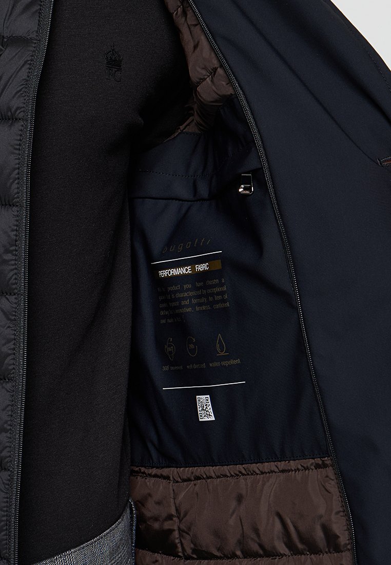 Schwarze Steppjacke mit brauner Innenauskleidung, ausgestattet mit einem Leistungsstoffetikett, Reißverschluss und Pflegehinweisen. Glatte Textur, wasserabweisend.