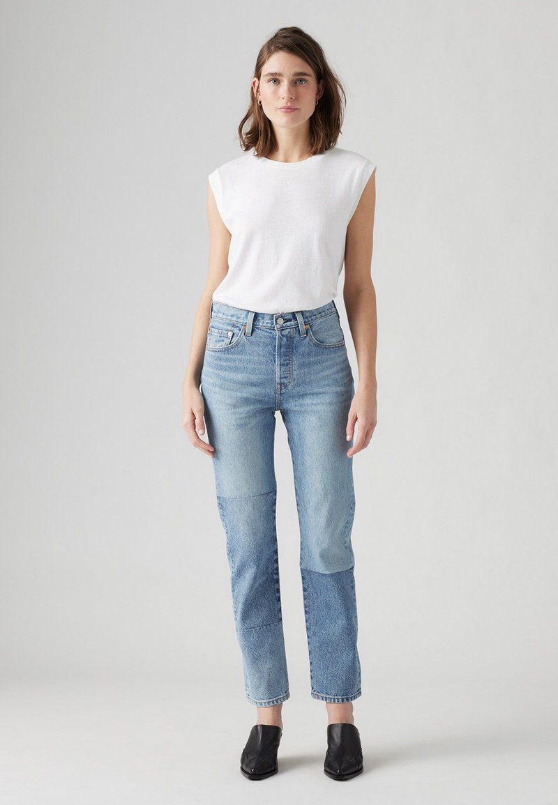 Top bianco senza maniche e jeans a toppa blu con vestibilità dritta. Scarpe col tacco nere. Tessuto in denim chiaro con tonalità variabili.