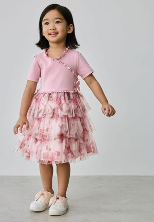 Young girl wearing a pink ruffled dress with floral tulle layers and white sneakers with large pink bows, standing on gray floor.