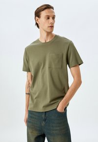 Oliven grøn bomulds t-shirt med korte ærmer, rund halsudskæring og en enkelt venstre brystlomme. Glat tekstur med minimale syninger.