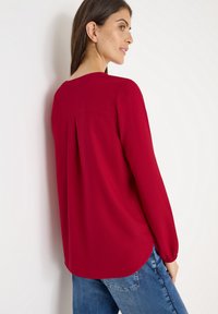 Rød bluse med lange ærmer, der har en rygfold og rund kant, lavet af letvægtigt stof. Sammen med blå denimjeans.