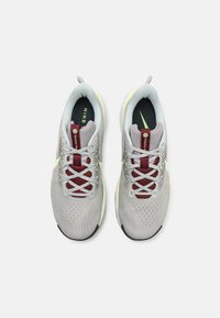 Un par de zapatillas de correr Nike de color gris claro con acentos amarillos pálidos y etiquetas de lengüeta en color marrón, vistas desde arriba sobre un fondo blanco.