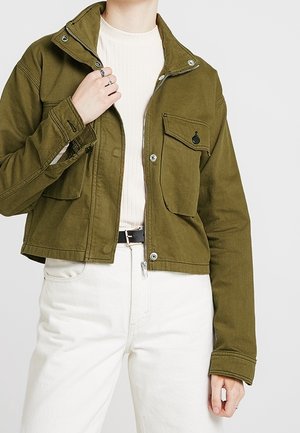 Persoon draagt een olijfgroen cropped jack met voorzakken, een wit geribd topje, witte hoge taillebroek en een zwarte riem.