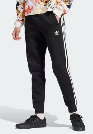 Nogi w czarnych spodniach dresowych Adidas z białymi paskami i czarnych sneakersach, jedna ręka w kieszeni, widoczna kolorowa wzorzysta bluza.