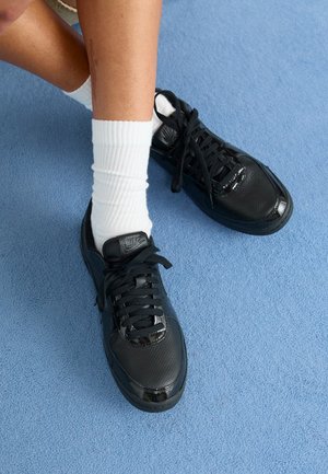 Nohy s bílými sportovními ponožkami v černých teniskách Nike, sedící na modrém koberci, s vázanými tkaničkami a zkříženýma nohama.