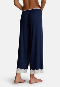 Vrouw in marineblauwe wijde broek met witte kantafwerking aan de zoom, staand op blote voeten met de rug naar de camera gekeerd.