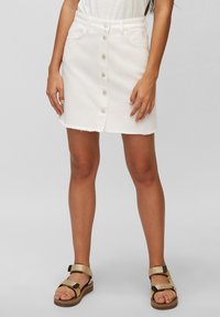 Minifalda de mezclilla blanca con dobladillo deshilachado, cierre frontal con botones y dos bolsillos. Combinada con sandalias marrones y doradas.