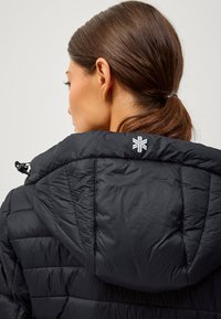 Sort pufferjakke med tekstureret finish, hætte og et geometrisk logo på ryggen. Detaljeret syning og quiltning er synlig.