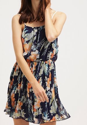 Vrouw draagt een mouwloze marineblauwe bloemenjurk met geplooide rok, houdt een hand in haar haar en laat de andere hand langs haar hangen.