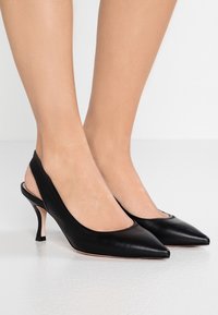 Des pieds portant des escarpins à talons kitten en cuir noir avec bout pointu, sur un sol et un fond blancs.