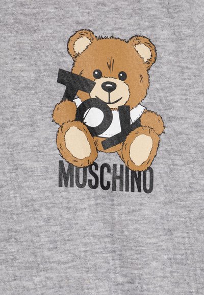 La tela de algodón gris presenta un oso de peluche marrón en dibujos animados con letras negras que dicen "Toy Moschino" debajo. El oso lleva una camiseta blanca y está sonriendo.