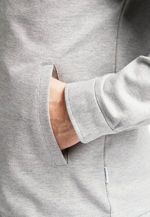 Hand in the pocket of a light grey long-sleeve sweatshirt with a small white fabric brand tag on the side seam.
