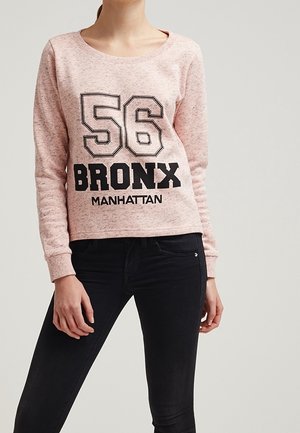 Helle rosa Sweatshirt mit Rundhalsausschnitt, mit einem schwarzen Grafikdruck: "56 BRONX MANHATTAN" in fetter Schrift. Kombiniert mit schwarzen Jeans.