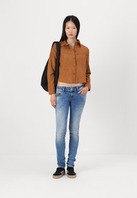 Chemise cropped en daim marron avec fermeture à boutons, jean skinny déchiré bleu clair, et baskets noires avec semelles beiges. Sac à dos noir sur l'épaule.