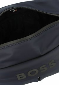 Bolsa de nailon azul marino con acabado texturizado, gran apertura con cremallera y logotipo "BOSS" en relieve en la parte frontal. Interior forrado en tela negra.