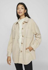 Giacca in misto lana beige con chiusura a bottoni, due tasche sul petto e vestibilità morbida, indossata sopra un maglione bianco con texture.