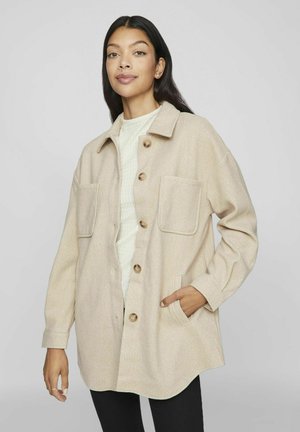 Giacca in misto lana beige con chiusura a bottoni, due tasche sul petto e vestibilità morbida, indossata sopra un maglione bianco con texture.