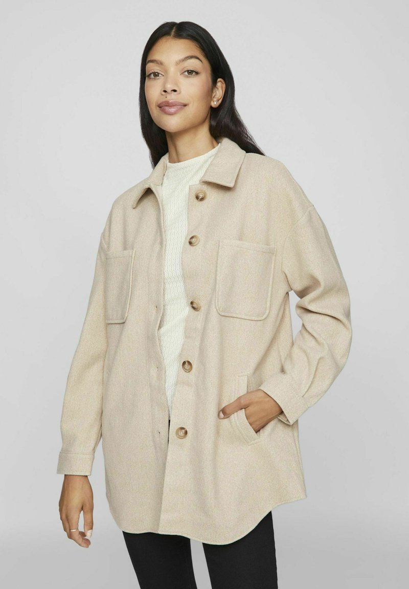 Giacca in misto lana beige con chiusura a bottoni, due tasche sul petto e vestibilità morbida, indossata sopra un maglione bianco con texture.