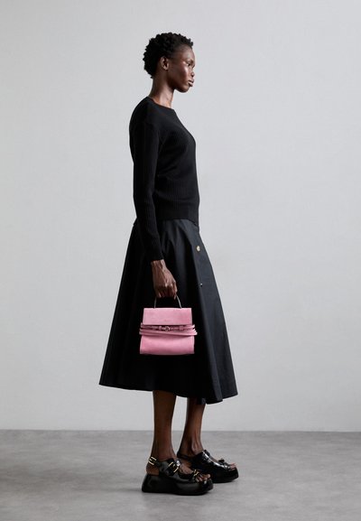 Melnā ribota džemperis apvienots ar melnu svārku. Modelis tur nelielu, strukturētu rozā zamša somiņu ar augšējo rokturi un sprādzes detaļu.