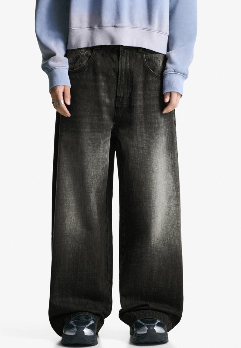 Persoană îmbrăcată în blugi negri oversized și decolorati, un hanorac bleu deschis, inel argintiu și adidași negri, stând pe un fundal alb.